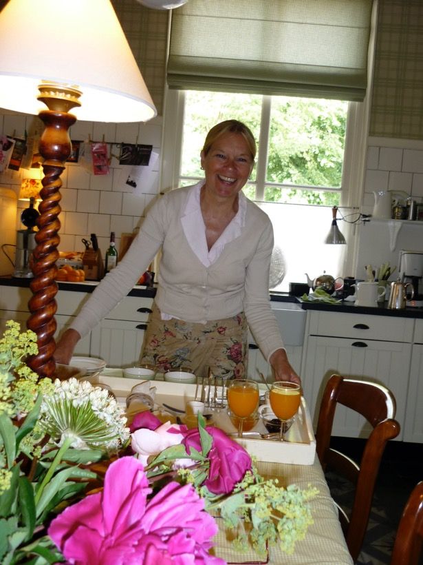 Antieke witte keuken met lachende gastvrouw in het midden. Gedekte tafel met ontbijt, glaasjes jus d'orange en een fuchsia-gekleurd bloemstuk op de voorgrond.