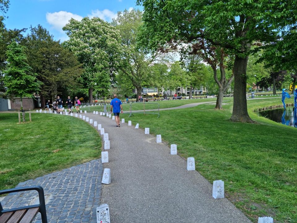 Deelnemers lopen over een parkpad in Woerden, gemarkeerd met lichtzakken tijdens de 24 Uur van Woerden, omgeven door groen, bomen en water op een zonnige dag.