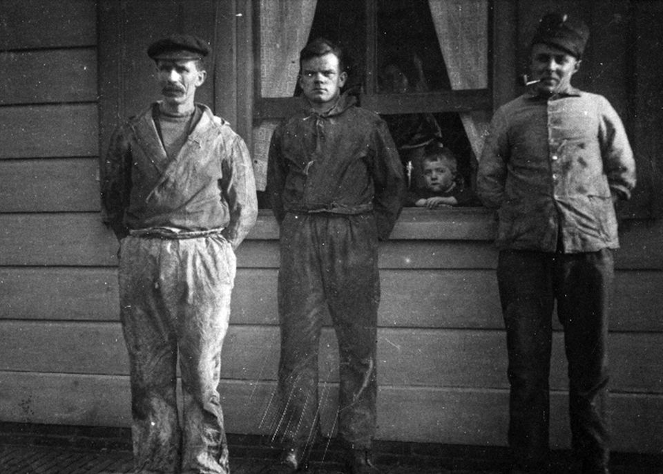 Zwart-wit foto van 3 mannen, waaronder fortwachter Linders (rechts) op Pampus. Op de achtergrond zie je het huis waar een kind het raam uitkijkt.