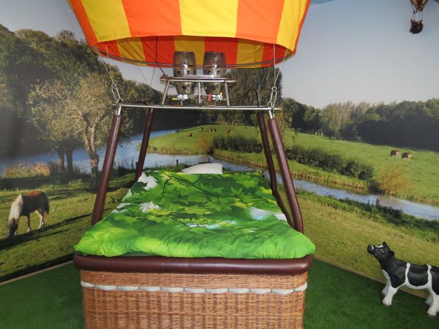 In een binnenruimte met fotobehang van een natuurgebied staat een mand van een luchtballon op de grond. Naast de ballon een beeld van een zwart bont kalfje.