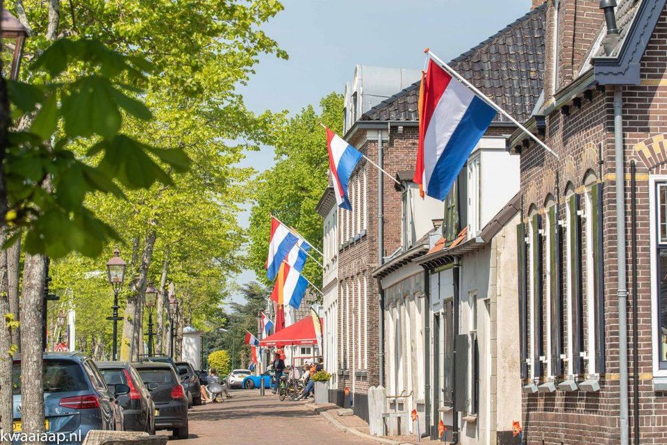 Dorpsstraat Kamerik, dorp in het Groene Hart bij Woerden, tijdens Koningsdag 2026 met Nederlandse vlaggen aan historische bakstenen gevels, geparkeerde auto’s en een feestelijke straat met oranje accenten en terras.