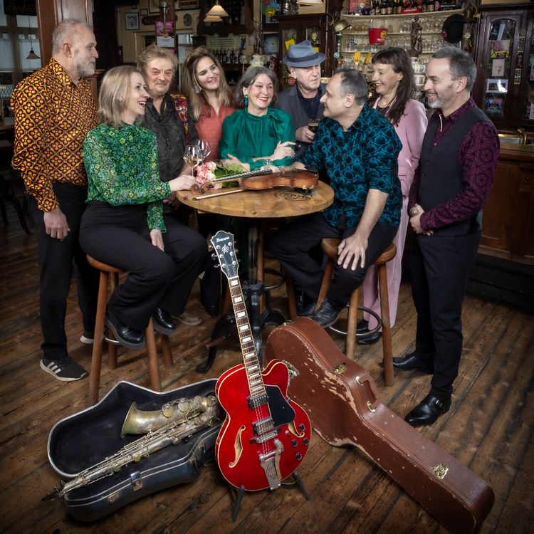 Foto van de tribute band zittend aan een hoge tafel in een café. De band bestaat uit vier vrouwen en vijf mannen. Op de voorgrond staat een gitaar en ligt een saxofoon in een opbergkoffer.
