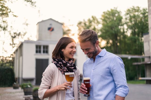 Een stel staat lachend voor de bierbrouwerij met een biertje in hun hand.