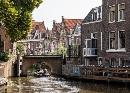 Een sloep vaart door een smalle gracht onder een lage brug, terwijl wandelaars de brug oversteken. Langs het water staan historische huizen met trapgevels en terrassen, omgeven door groen, in de binnenstad van Oudewater.