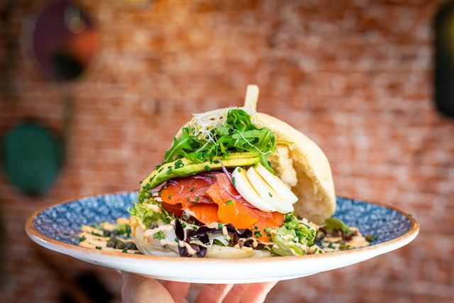 Close-up van een broodje, bereid door Broodje Midas uit Woerden, met gerookte zalm, avocado, gekookt ei, rucola en kiemen, geserveerd op een rustiek bord tegen een bakstenen achtergrond.