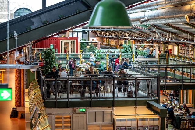 Bezoekers studeren en werken aan tafels op een tussenverdieping in een moderne bibliotheek, met balustrades, industriële verlichting en zicht op lagere verdiepingen met boekenkasten.