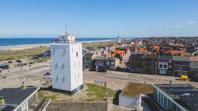 Dronebeeld van de witte vuurtoren in Katwijk aan Zee, met op de achtergrond huizen en de Boulevard, een duinenrij, het strand en de Noordzee.