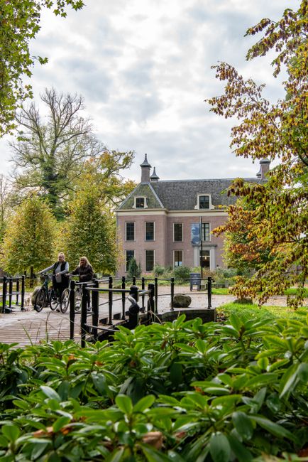 Twee vrouwen lopen met de fiets in de hand over de brug voor Landhuis Oud Amelisweerd