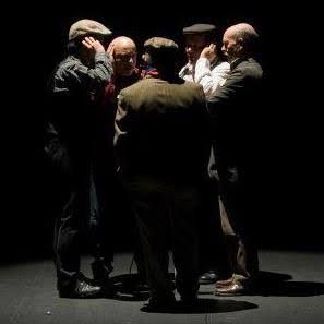 Donkere foto van vijf mannen, sommige met pet, sommige kaal, die in een kring staan met hand aan hun oor.