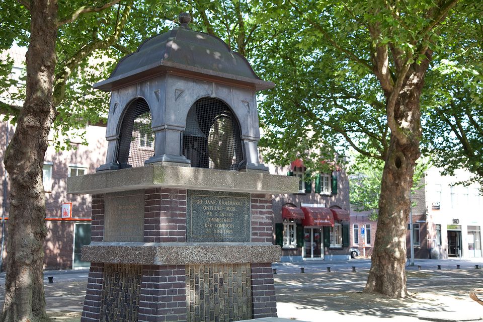 De Kaasbel op het Kerkplein in Woerden, een monument ter ere van 100 jaar kaasmarkt, omringd door bomen en historische panden.