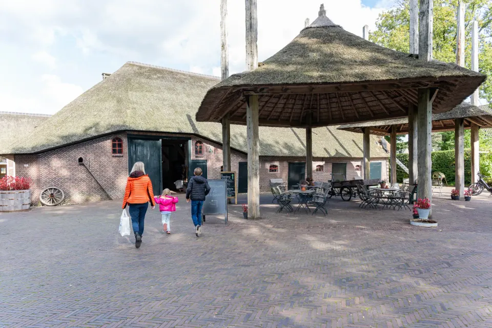 Een volwassene en twee kinderen lopen het erf van Museumboerderij Het Gagelgat op, met de rietgedekte boerderij op de achtergrond.