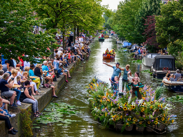 Veel mensen die toekijken terwijl boten, versierd met bloemen door de grachten van Delft varen