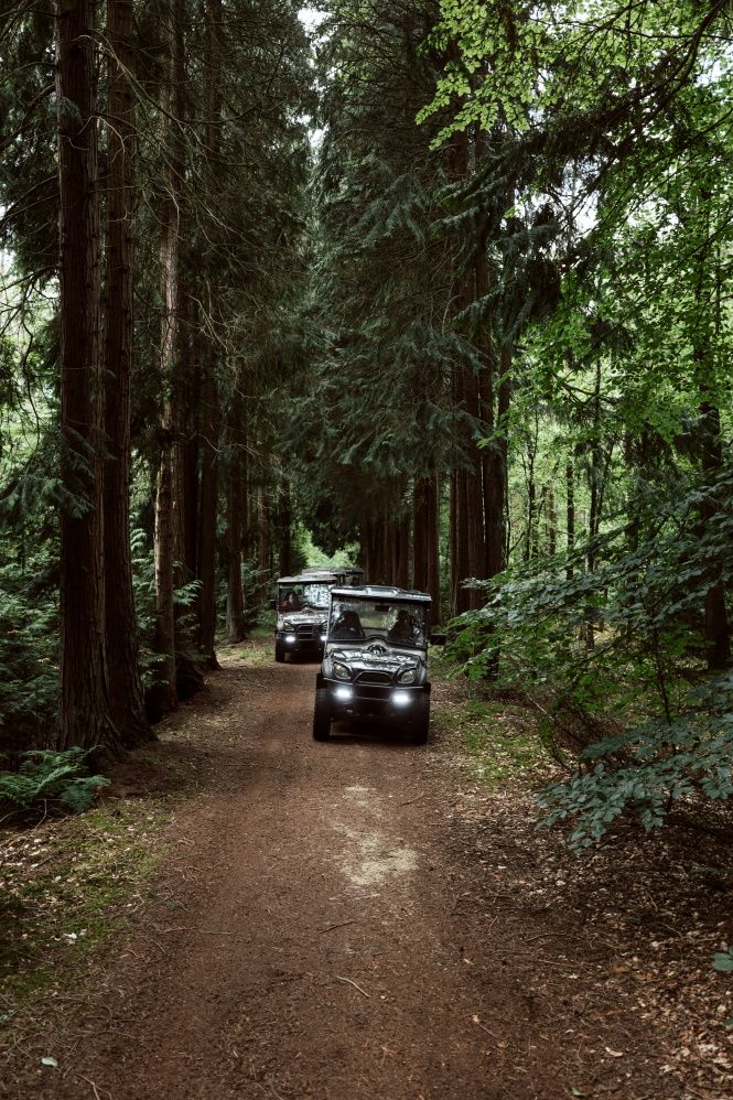 2 elektrische jeeps rijden achter elkaar over een onverhard pad tussen hoge groene naaldbomen door.