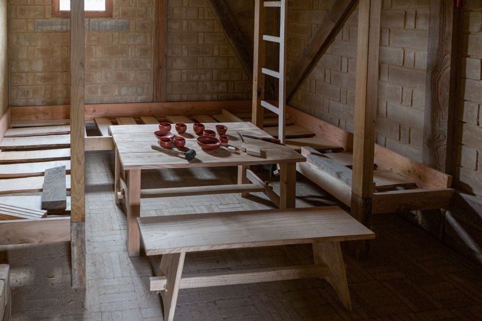 Houten banken en tafels in de soldatenbarak, met op tafel aardewerk drinkgerei.