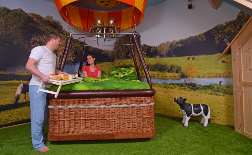In een binnenruimte staat een mand van een luchtballon op de grond. Voor de mand staat een man met een dienblad.  Achter de ballon staat een mevrouw en naast de ballon een beeld van een zwart bont kalfje.