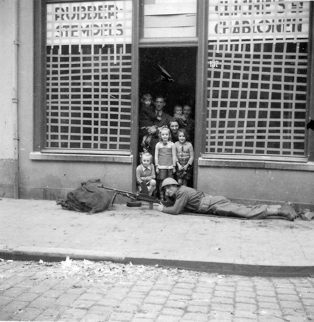 Een soldaat van het Welsh Regiment tijdens de bevrijding van ’s-Hertogenbosch, 28 oktober 1944. De foto is door J. de Zeeuw gemaakt voor de deur van Klaassen, zaak in rubber, stempels, clichés en sjablonen, Hinthamerstraat 137. Ondanks dat de ramen zijn afgeplakt met papierstroken ter bescherming tegen glasscherven, staat de familie Klaassen in de deuropening.