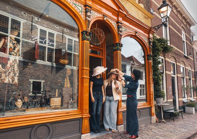 Hoedenatelier Beau Chapeau in Oudewater, Groene Hart. Ambachtelijke hoedenwinkel met historische gevel waar een hoedenmaakster twee bezoekers helpt met het passen van stijlvolle hoeden voor de entree in de sfeervolle binnenstad.