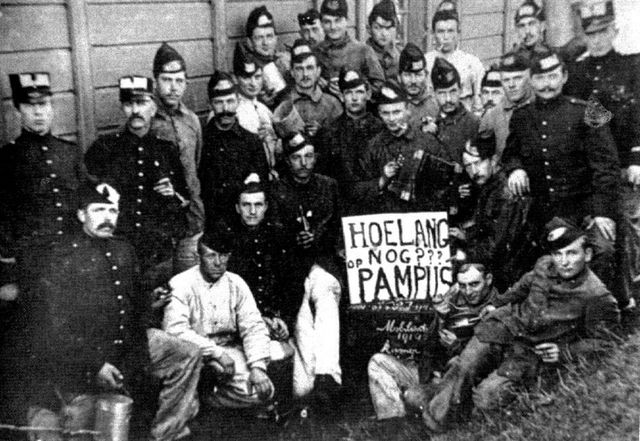 Een grote groep soldaten vragen zich af hoe lang ze nog op Pampus moeten verblijven. Ze kijken allemaal recht de camera in terwijl ze een bord vasthouden met die vraag.