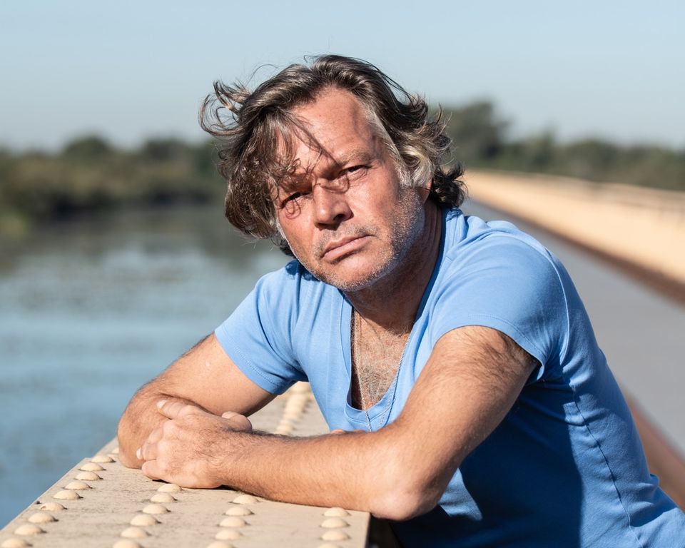 Portret van Serge van Duijnhoven die leunt op de rand van de brug. Op de achtergrond zijn een rustige rivier en een brug te zien bij helder daglicht.