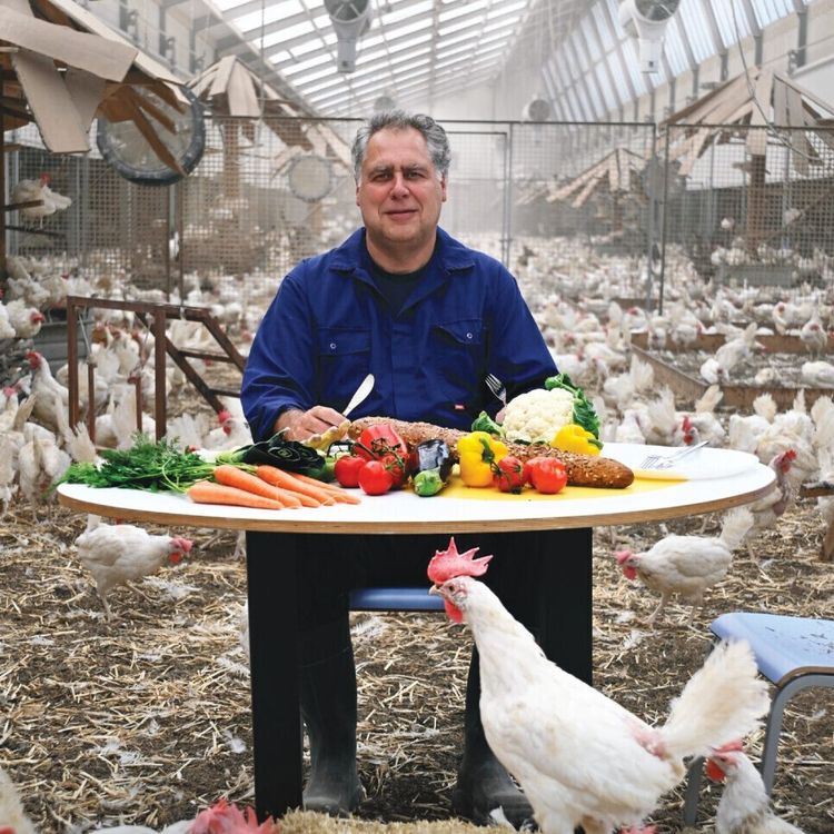 Foto van een boer in blauwe overall in een kippenstal achter een ronde tafel met kleurrijke groenten.
