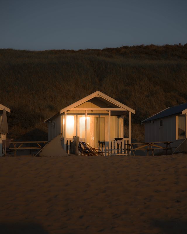 Foto van een strandhuisje in Katwijk aan Zee, tegen de duinenrij aan, met reflectie van ondergaande zon in de ramen van het middelste huisje, tussen twee andere vrijstaande huisjes in.