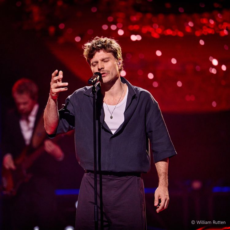 Vrij donkere foto van Charl Delemare achter een microfoon op standaard met flonkerende roze en rode lichtjes achter hem.