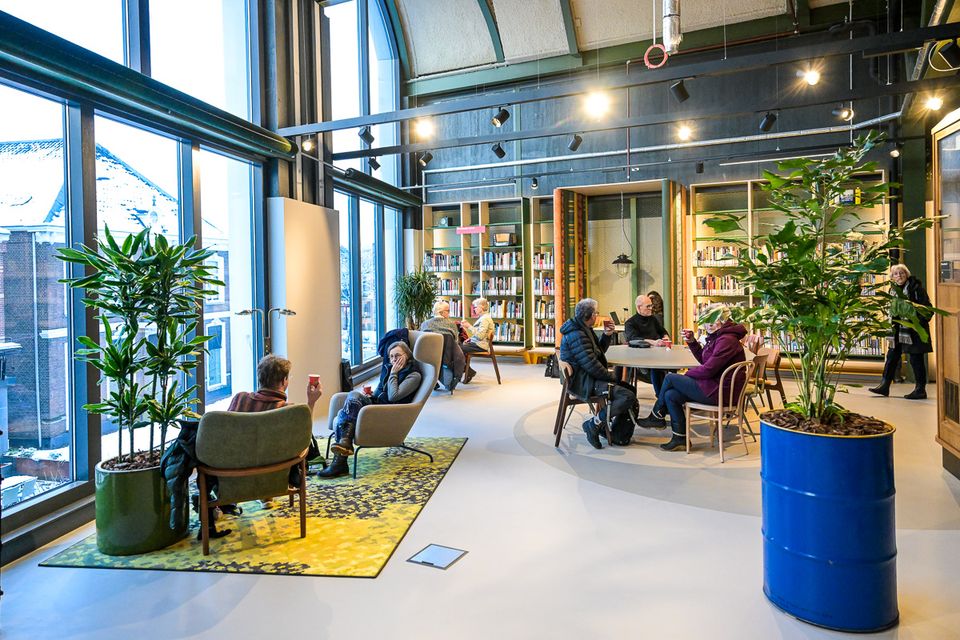 Lichte leesruimte in een bibliotheek met grote ramen, planten en boekenkasten. Bezoekers zitten in stoelen en aan tafels, praten met elkaar en drinken koffie in een informele, rustige omgeving.