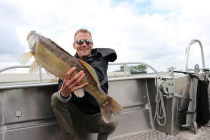 Een man met een zonnebril houdt glimlachend een grote vis omhoog, terwijl die gehurkt op een boot op het water zit.