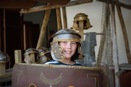 Een jongen die een romeinse helm op heeft en een schild vast houd met daarachter nog meer romeinse leger spullen.