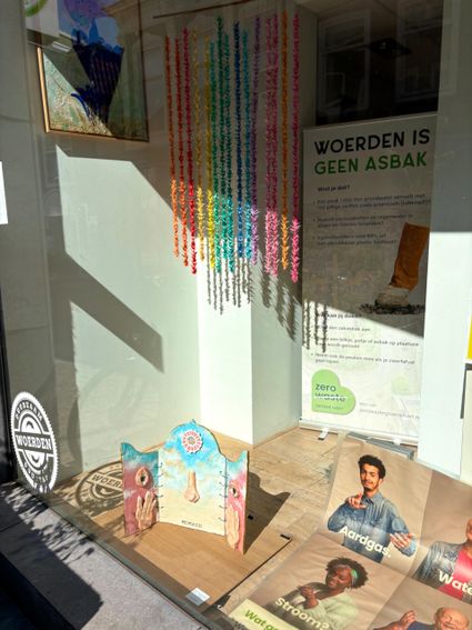 Etalage in Woerden met kleurrijke kunstinstallatie en campagneposter ‘Woerden is geen asbak’ van Zero Waste Groene Hart tegen zwerfafval en sigarettenpeuken.