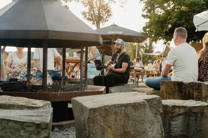 Man speelt gitaar en zingt bij een vuurkorf op een terras, terwijl bezoekers op banken en aan tafels luisteren en drinken. Informele buitensfeer met parasols en zomers licht, wat duidt op een kleinschalig live optreden.