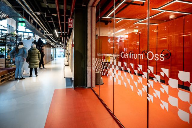 Gang in een modern gebouw met bezoekers die lopen langs een glazen wand met de tekst “Historisch Informatie Centrum Oss”, uitgevoerd in oranje tinten en industriële verlichting.