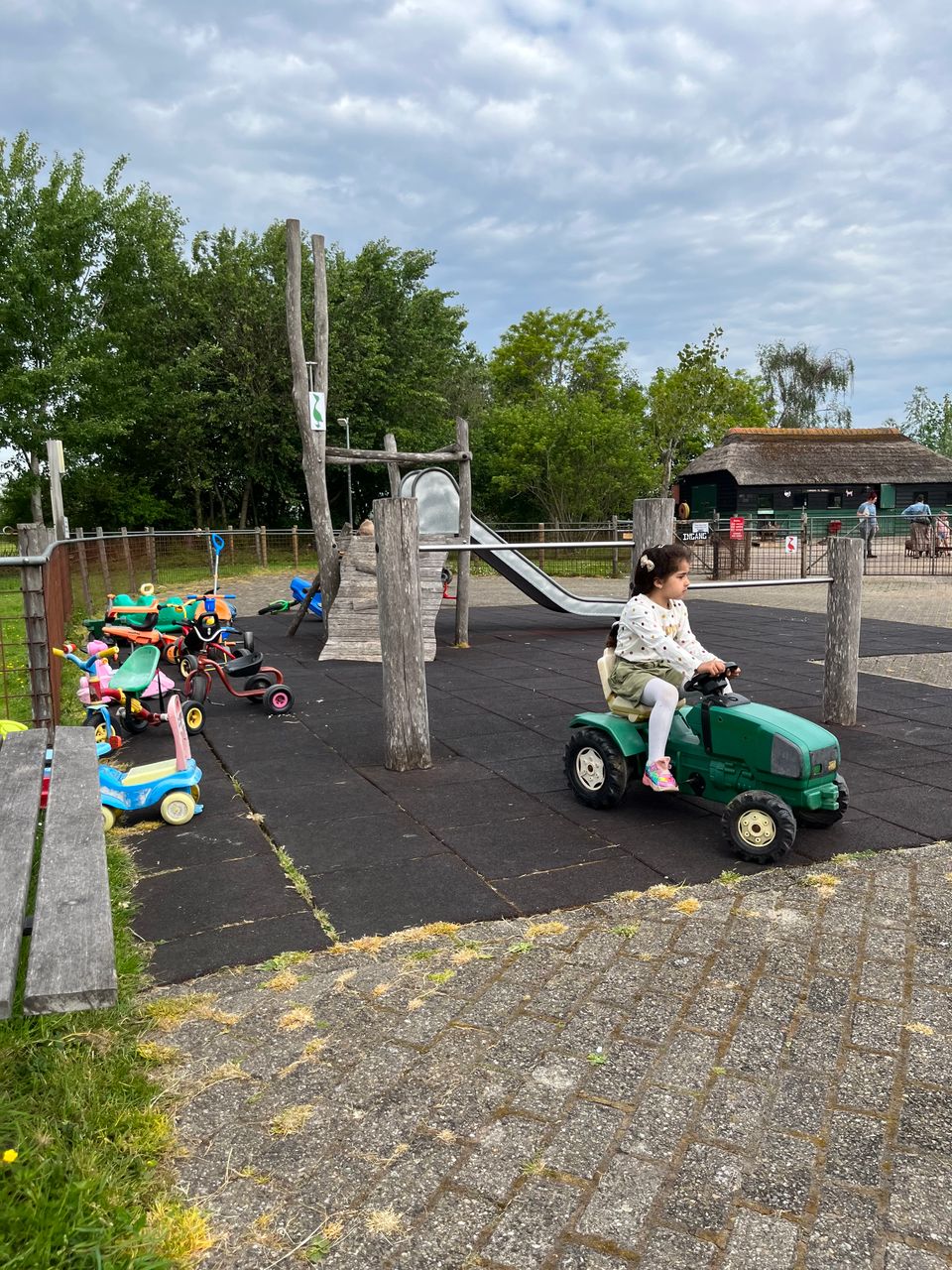 Kind speelt op een groene traptractor in de speeltuin van de kinderboerderij, met glijbaan en speelgoed op de achtergrond.