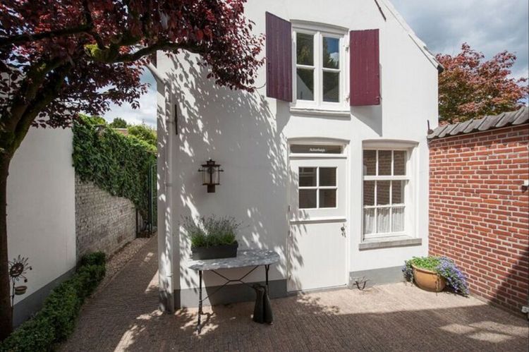 Wit geveltje met witte voordeur, rode luiken bij een bovenraampje, tafeltje met lavendel naast de voordeur. Op de voorgrond donkere bladeren van een boom.