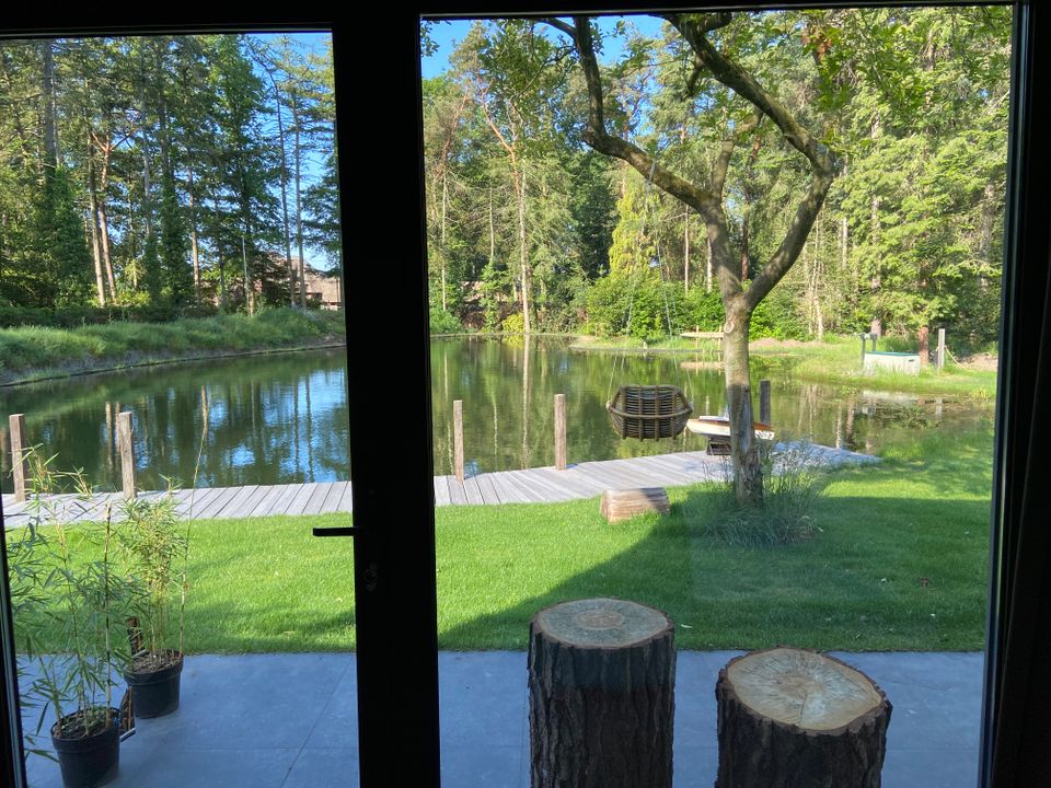 Uitzicht door openslaande deuren naar de vijver met houten vlonder en groen grasveld met daarop een boom met hangstoel. Bosrijk.