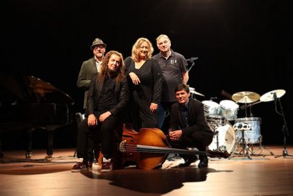 Vijf muzikanten poseren op een podium met instrumenten, waaronder een contrabas, piano en drumstel. Ze dragen donkere kleding en kijken richting de camera. De setting en belichting duiden op een bandfoto voorafgaand aan een optreden.