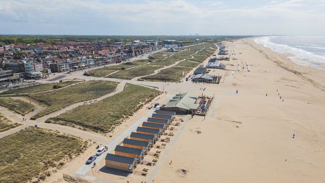 Dronebeeld van het strand, duinen en de Boulevard in Katwijk aan Zee. Met strandvakantiehuisjes, strandpaviljoens, het zandstrand, ze zee, een duinenrij en het centrum van Katwijk aan Zee.