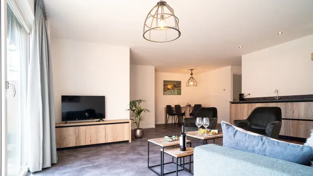 Zitkamer met lichthouten kasten en tafeltjes. TV en bank, met aan de rechterkant een keukenblok en links openslaande deuren.