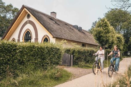Fietsroute langs een karakteristieke boerderij in het Groene Hart, waar twee vrouwen over een landelijk pad fietsen langs een rietgedekte woning en groene hagen. Foto: Studio Chanou.