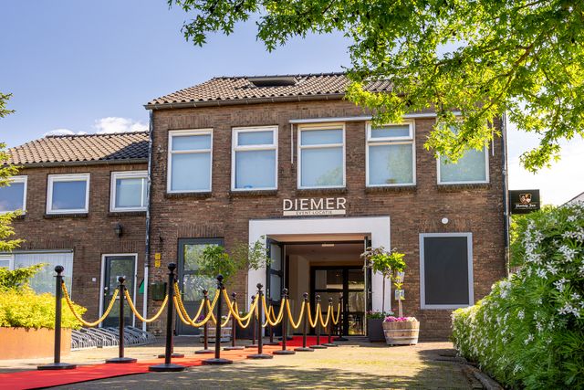 Voorgevel van Diemer Event Locatie in Oudewater met rode loper, goudkleurige afzetkoorden en bloembakken, onder een strakblauwe lucht en groene bomen.