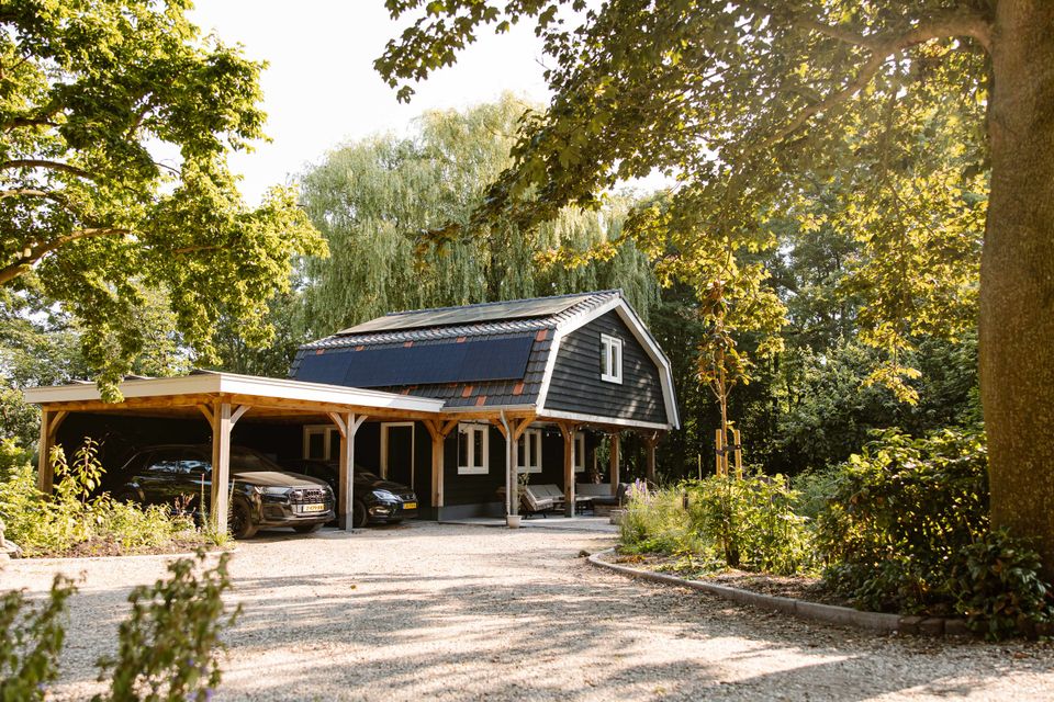 Landelijke vergaderlocatie Esti Yoga in Lopik, Groene Hart, met donkerhouten gebouw, overdekte veranda, carport en groene tuin omringd door bomen.