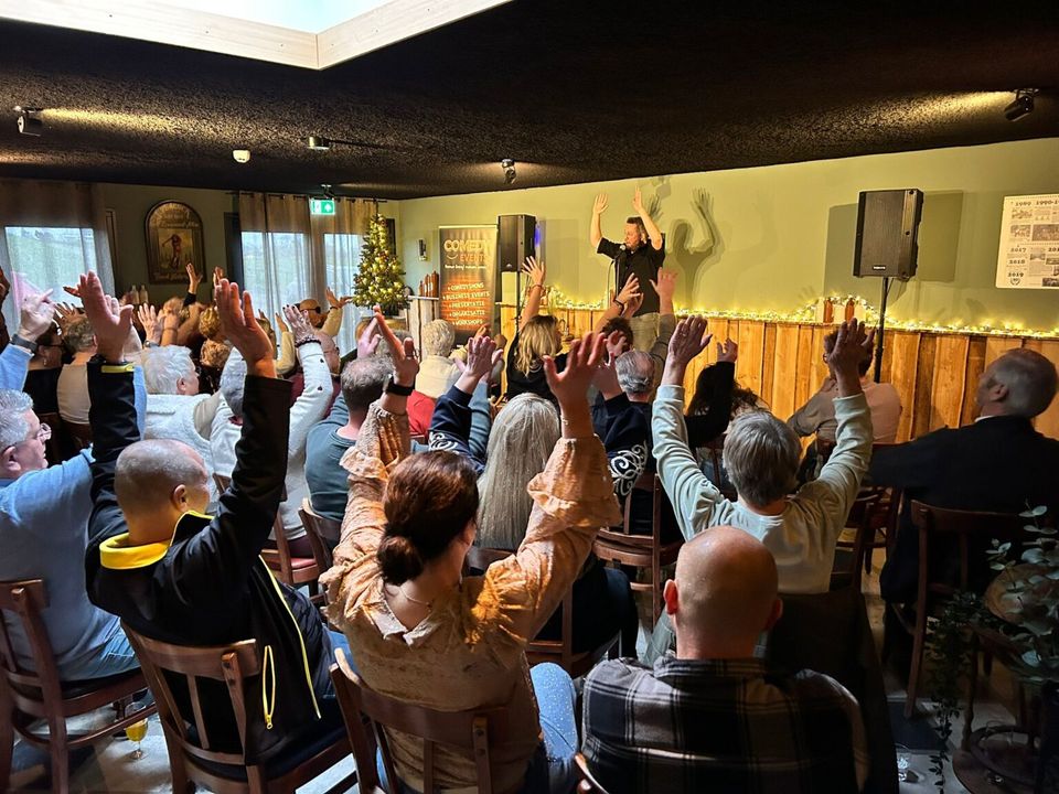 Zittend publiek in een kleine zaal steekt massaal de handen omhoog richting een spreker op het podium. Informele setting met stoelen, kerstverlichting en geluidsinstallatie, wat wijst op een interactief optreden of voorstelling.