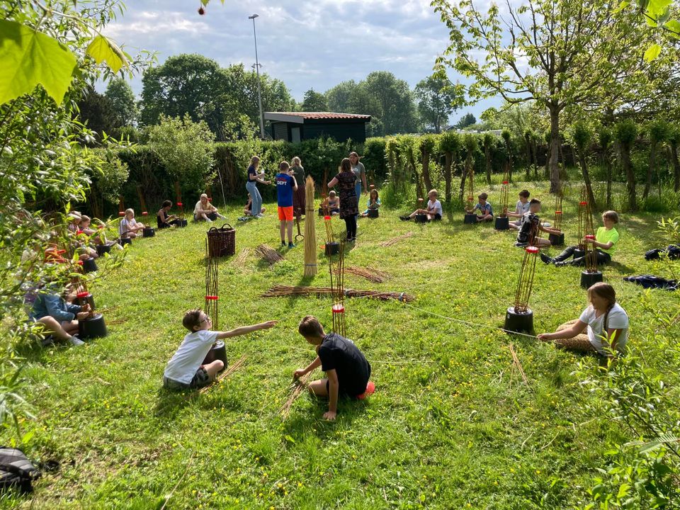 Workshop manden maken De Broedkorf in Oudewater, Groene Hart, groep kinderen en volwassenen vlechten met wilgentakken in het gras tijdens ambachtelijke buitenactiviteit met natuurlijke materialen.