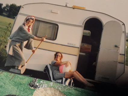 Foto van een witte caravan met 2 toneelspeelsters verkleed als haai in een bootje en als matroos met een roze strandbal.
