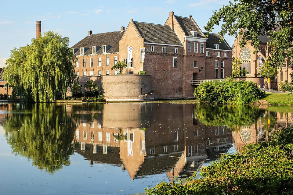 Zicht op kasteel Woerden, omringende groene bomen en struiken op een zonnige dag, inclusief water weerspiegeling.