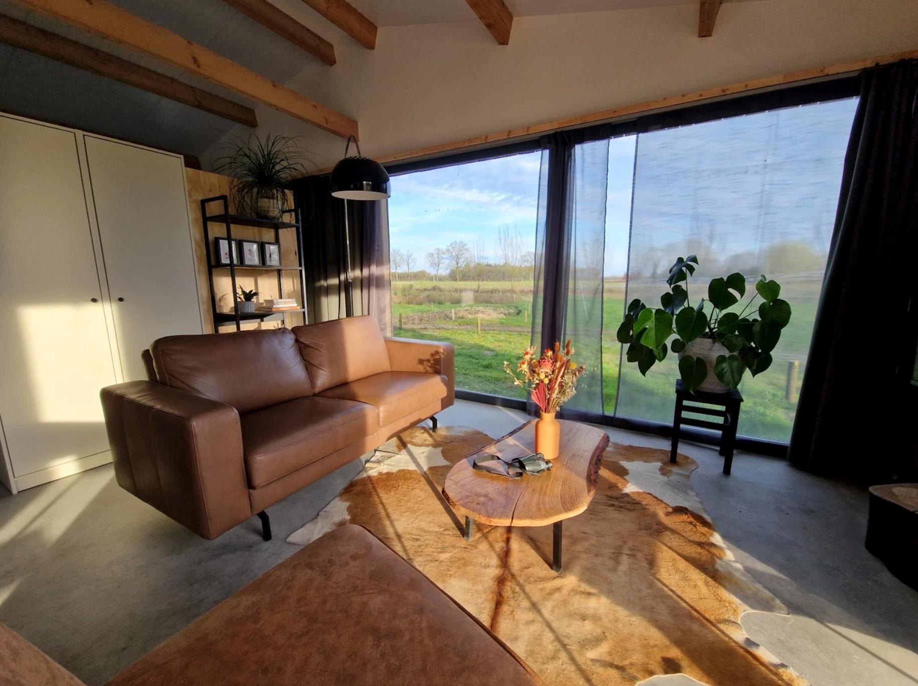 Woonkamer met bruine bank, koeienvel op de vloer, een houten salontafeltje en uitzicht op het platteland.