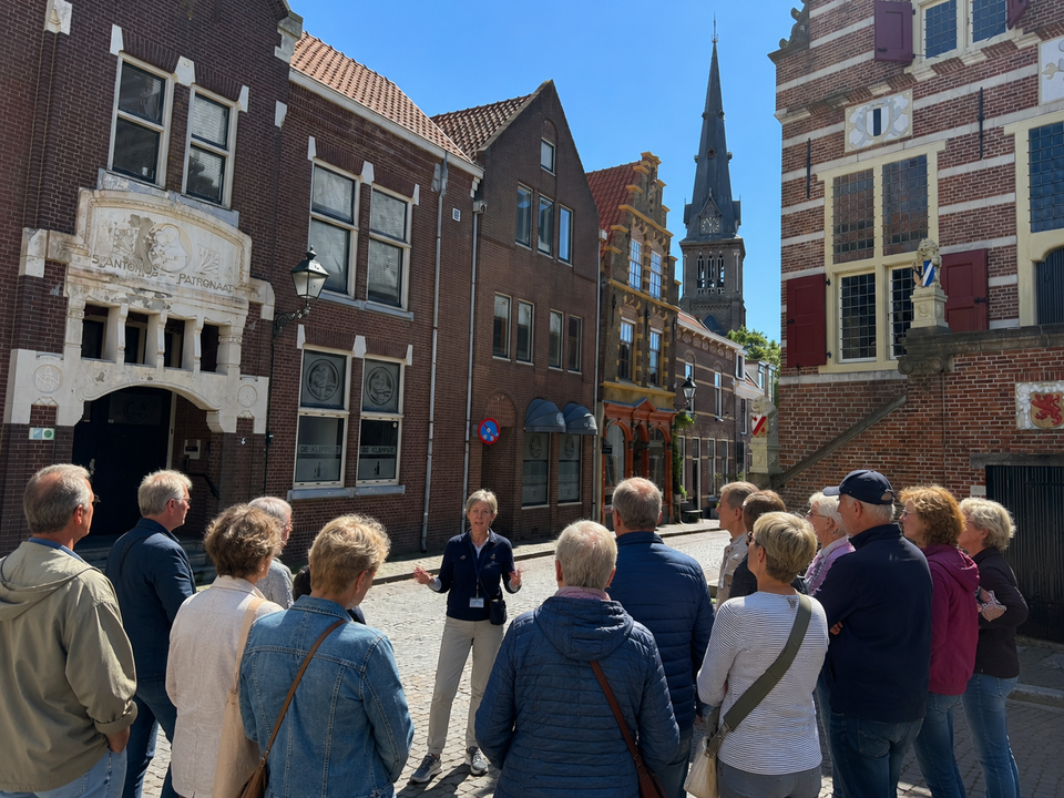 Een gids vertelt een groep bezoekers over de geschiedenis van Oudewater tijdens een stadswandeling op de Markt, met het historische stadhuis en de Sint-Michaëlskerk op de achtergrond.