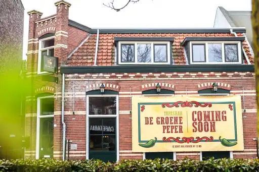 Vooraanzicht van een laag, historisch pand met bakstenen gevel en zadeldak. Rechts hangt een groot bord met de tekst “De Groene Peper – Coming Soon”. Links is een glazen deur met raam. Voor het gebouw staat groen.