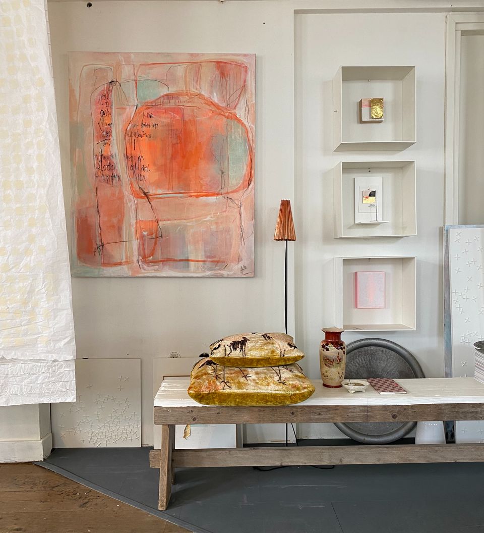 Interieur met kunstwerken: een groot abstract schilderij in zachte roze- en oranje tinten aan de muur, kleine kunstobjecten in nisjes en een houten tafel met decoratieve kussens, vaas en lamp.