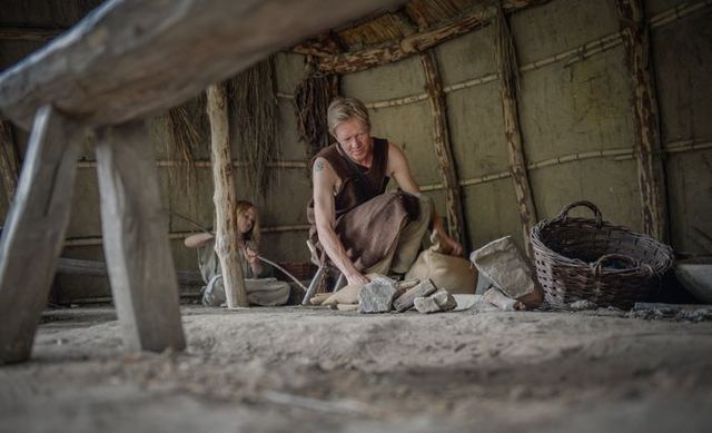 Man en kind in prehistorische kleding werken met stenen en gereedschappen in een gereconstrueerde hut uit de steentijd in Archeon, Groene Hart.